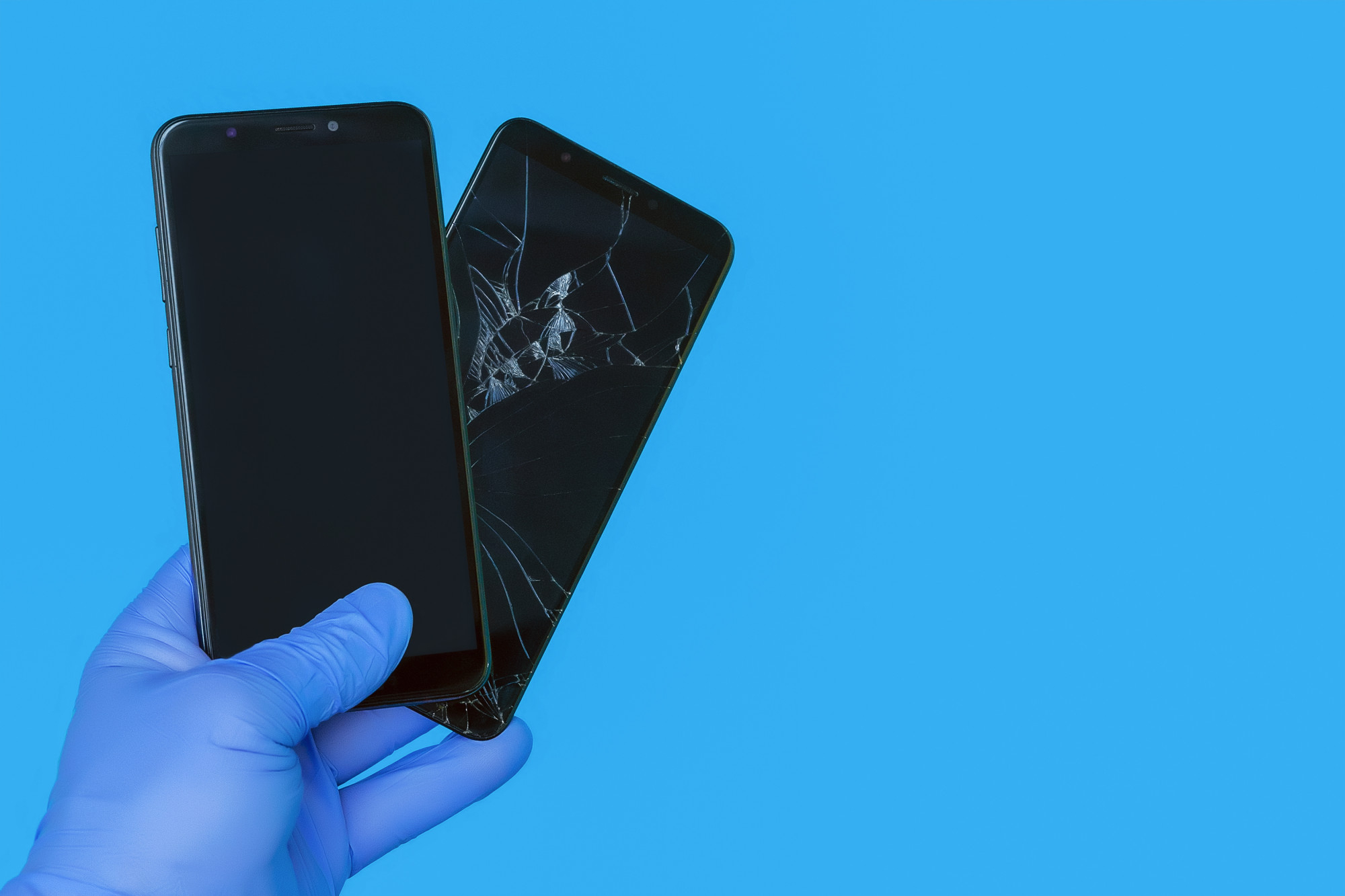 A hand holding two smartphones against a blue background, one with a cracked screen, suggesting device insurance fraud.