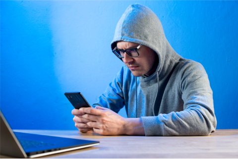 Man in hoodie using smartphone with laptop on desk against blue background.