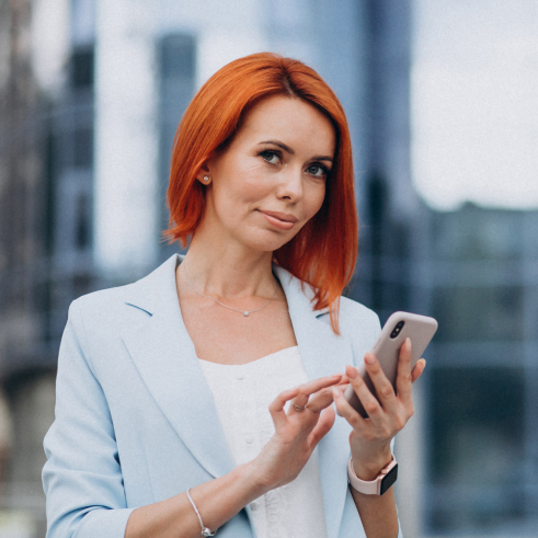 A woman with red hair holding a smartphone in an urban setting.
