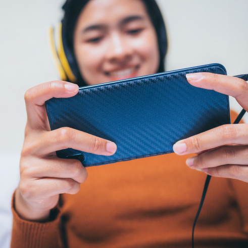 Person in an orange sweater holding a smartphone horizontally with headphones on, displaying a cheerful expression possibly engaging in entertainment or a video call.