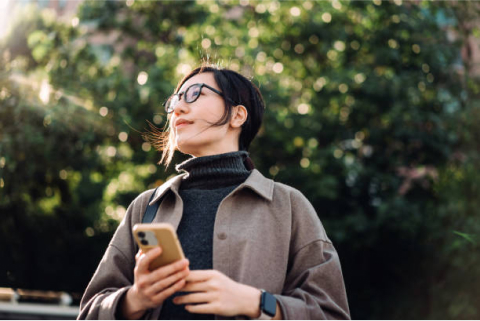 Woman holding a smartphone looks thoughtfully into the distance outdoors.