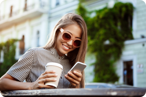 Mujer joven sonriendo a su teléfono mientras sostiene una taza de café al aire libre.